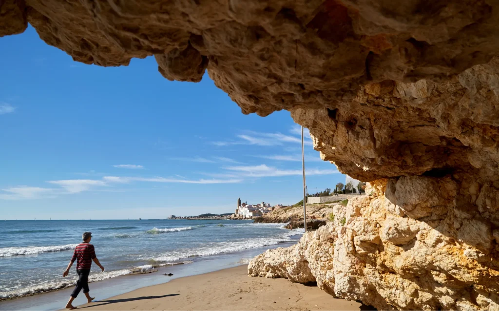 Persona contemplando la costa de Sitges desde bajo un arco de roca natural con el mar y la iglesia al fondo.