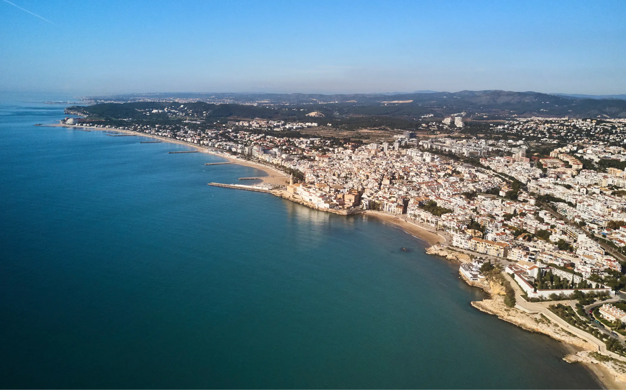 Aerial view of the entire coastline of Sitges with the beaches, the church, the harbor, and the Garraf mountain range.