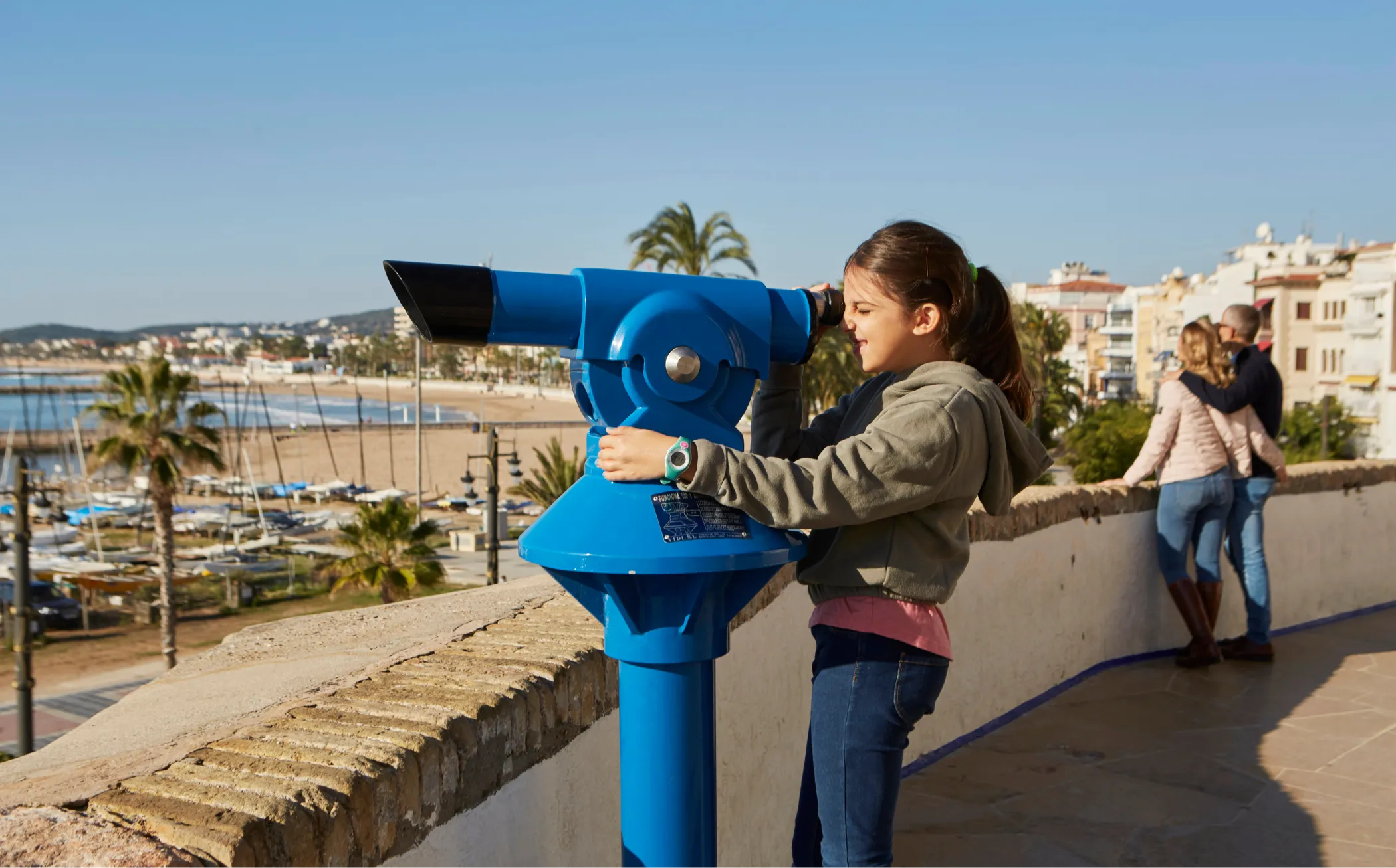 Nena mirant per un telescopi de monedes blau amb vistes al port i a les platges de Sitges.