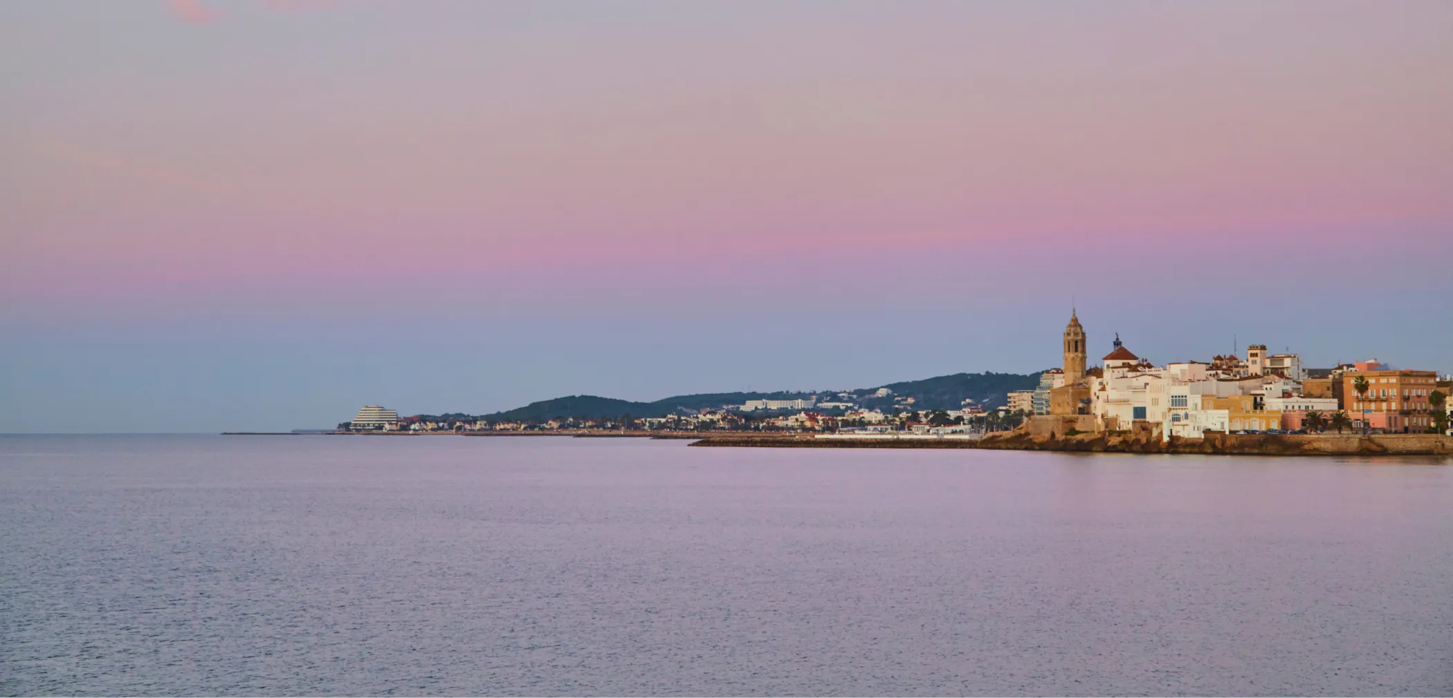 Panoràmica de la vila de Sitges al crepuscle amb el cel rosa-lila reflectit al mar tranquil.