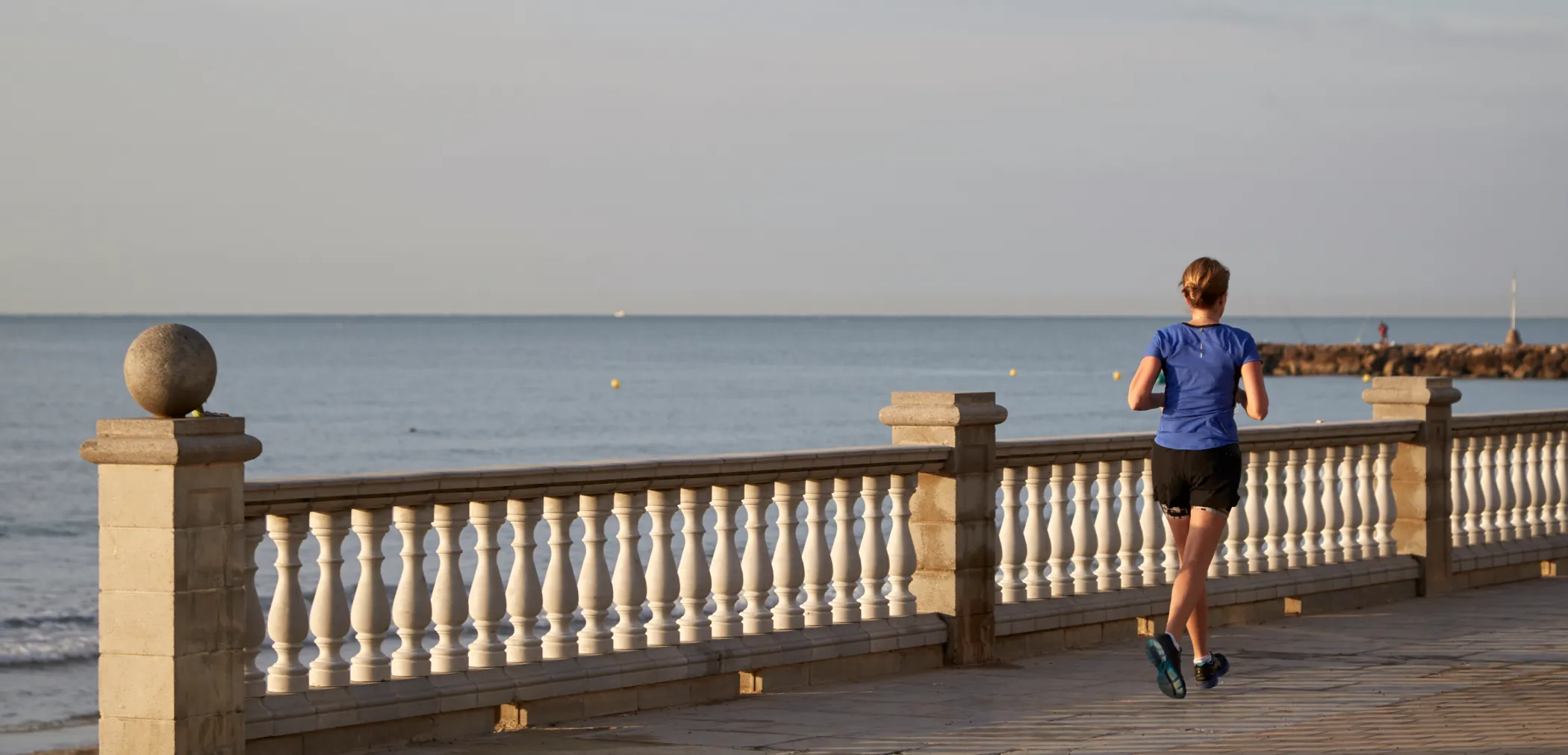 Corredora fent footing pel passeig marítim de Sitges, amb una balustrada de pedra i el mar de fons.