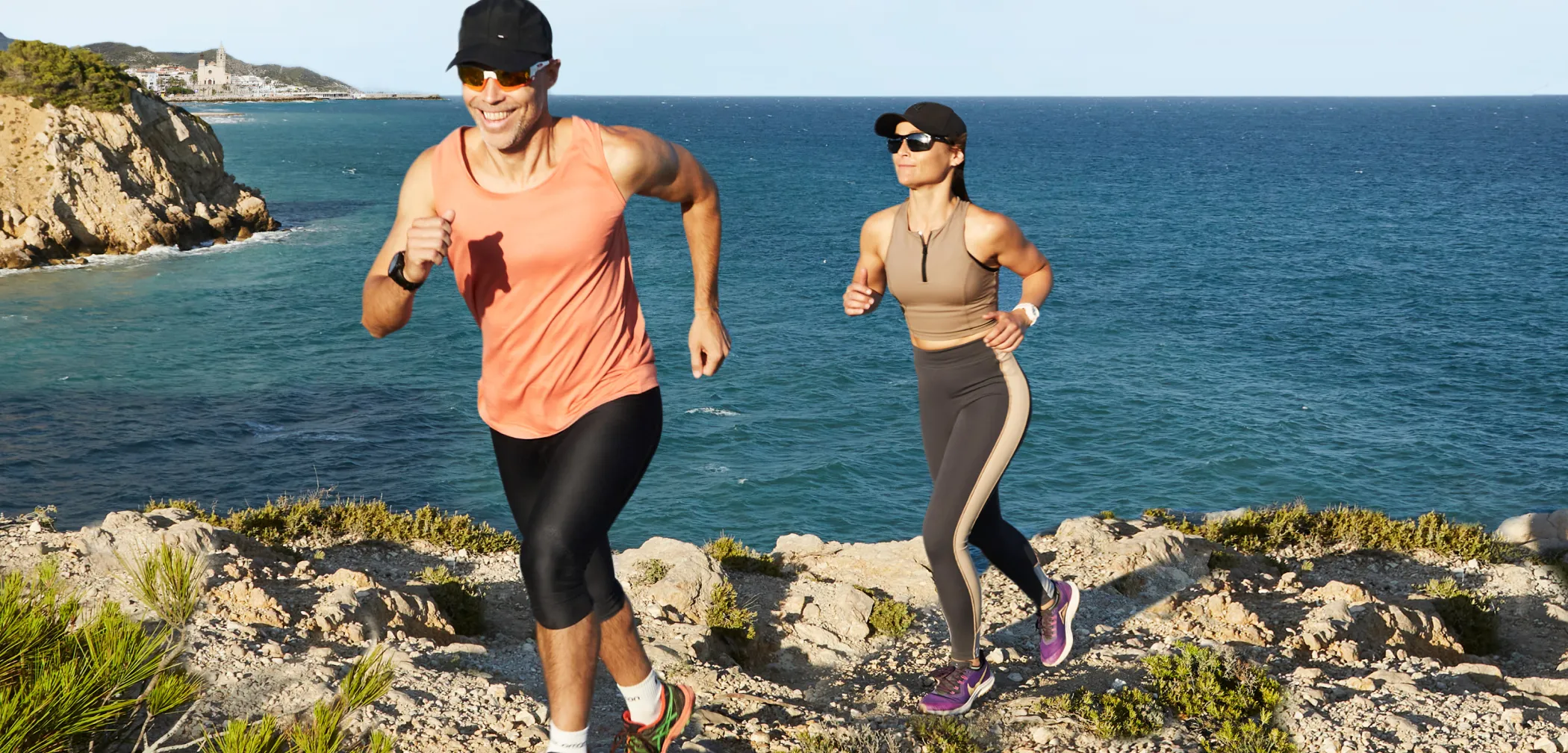 Dos corredors en forma esportiva corrent amb la costa de Sitges i el mar Mediterrani al fons.