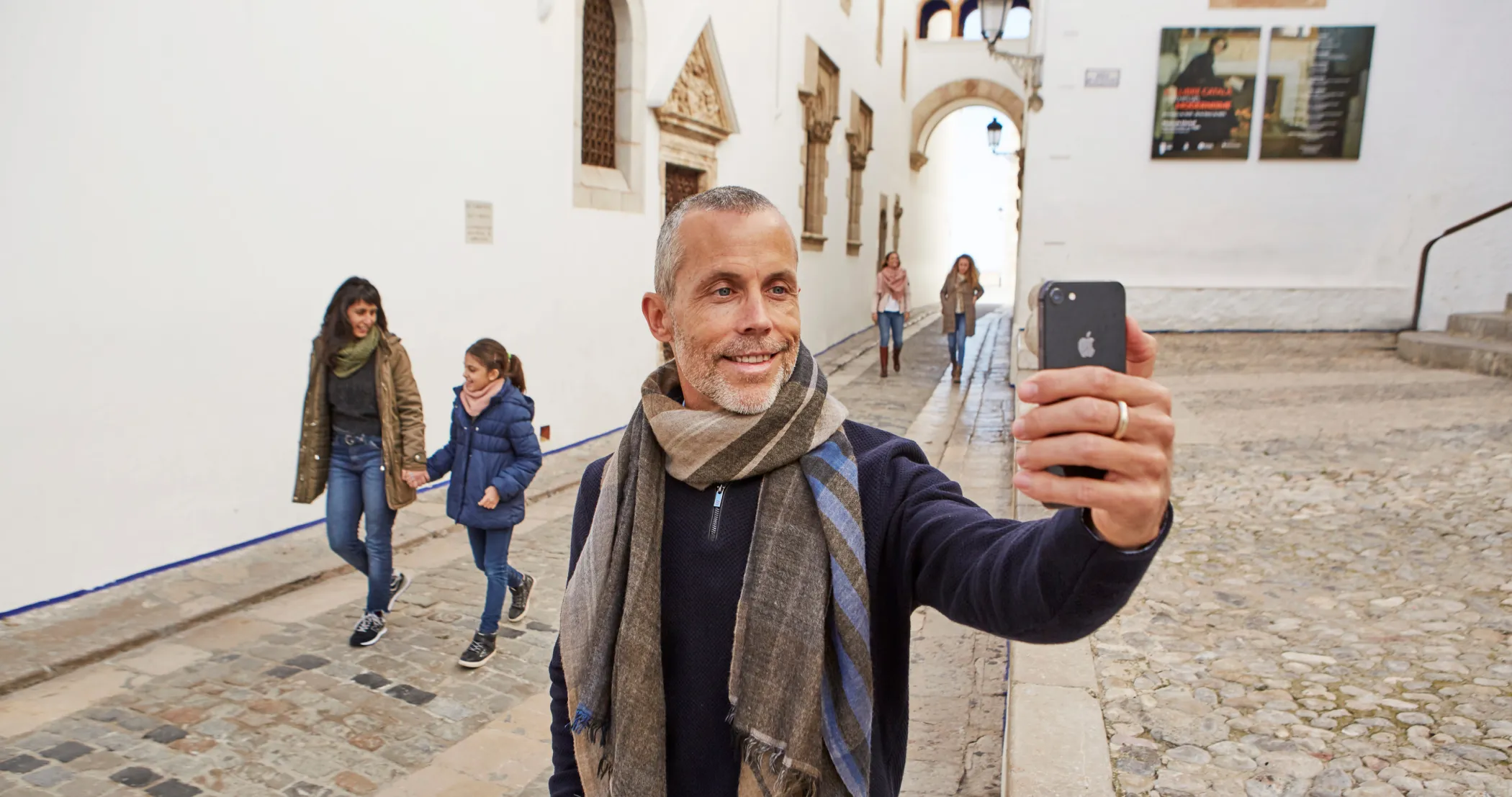 Home fent-se un selfie en un carrer empedrat del centre històric de Sitges amb una família al fons.