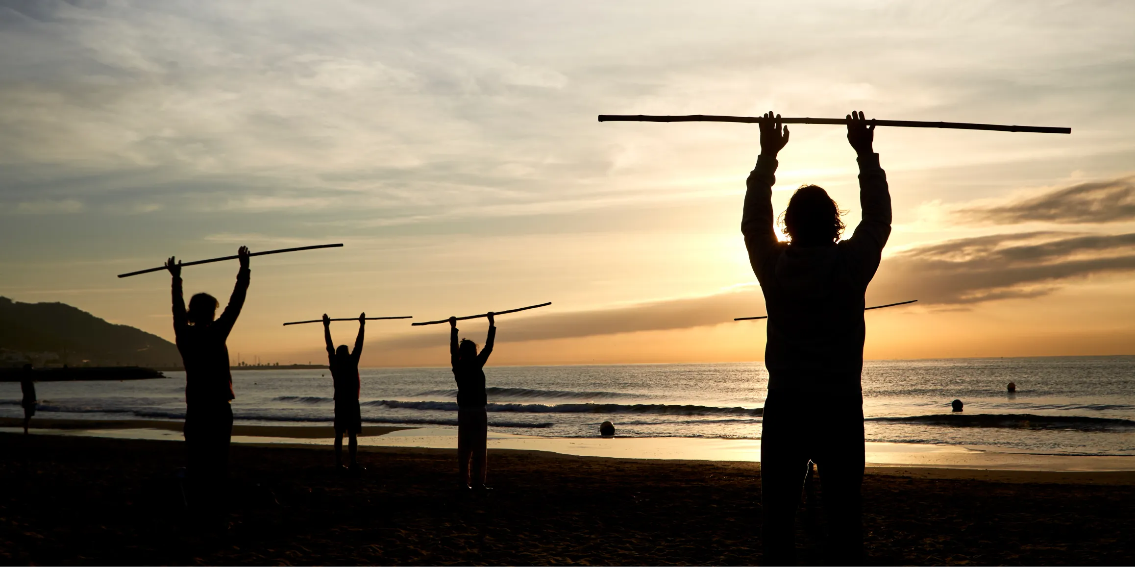 Three silhouettes of people exercising on the beach at sunset, holding horizontal sticks overhead.