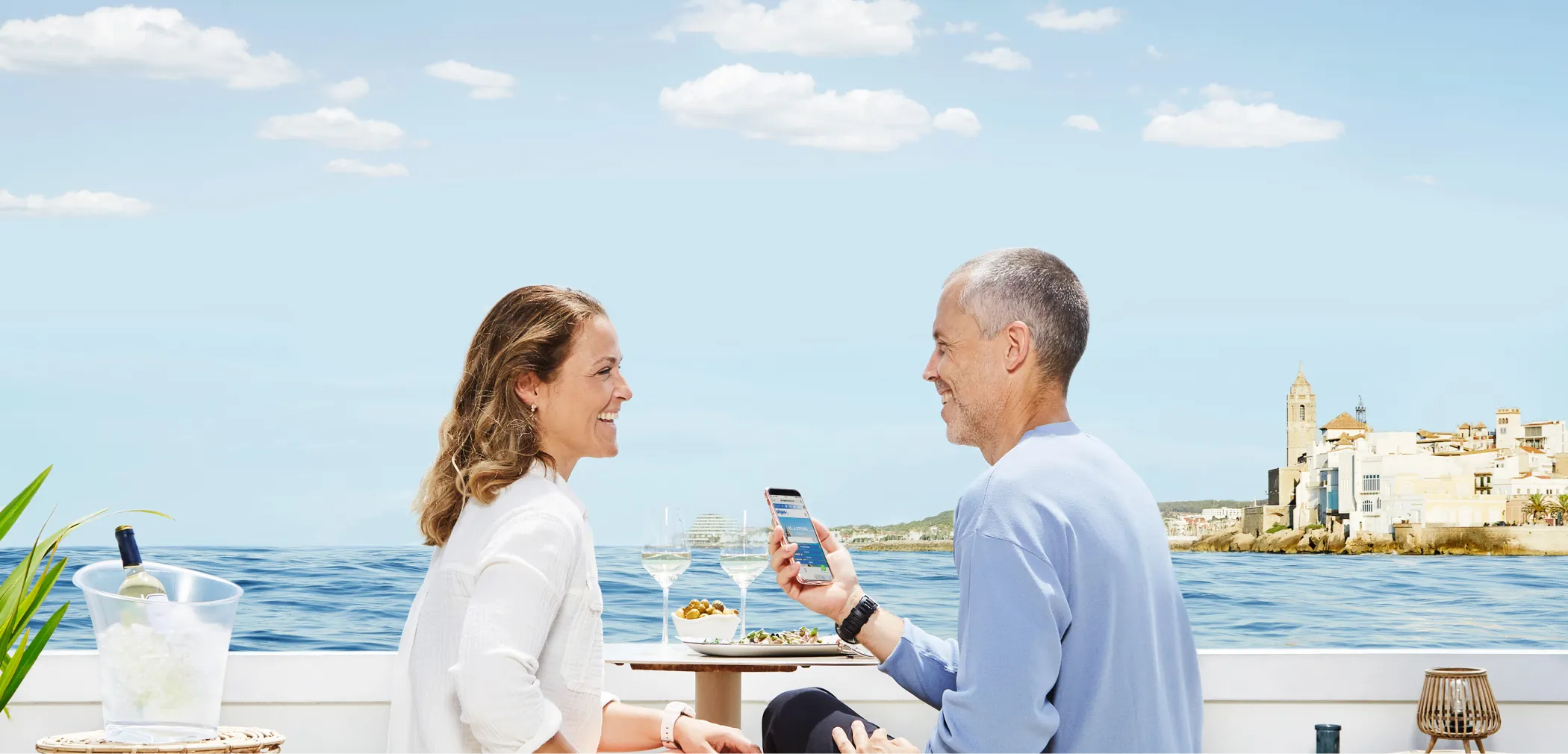 Two people chatting on a terrace overlooking the sea with the Church of Sitges in the background.