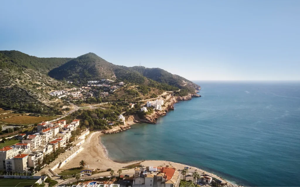 Vista aérea de la costa del Garraf con colinas verdes, acantilados y casas blancas junto al mar.