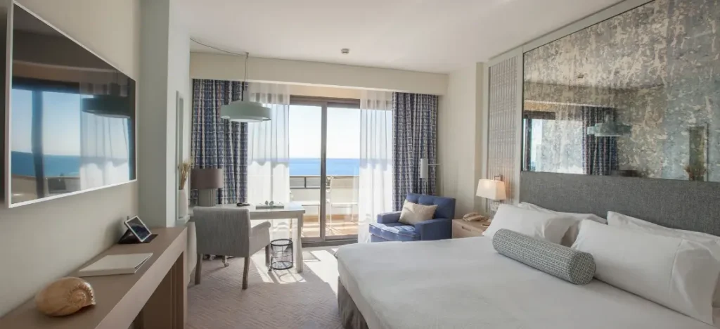 Habitación de hotel de lujo con cama grande, zona de asiento y balcón con vistas al mar Mediterráneo.
