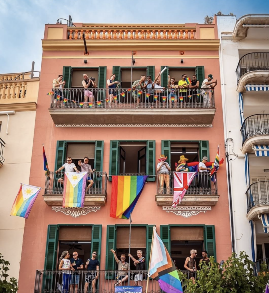 Façana amb balcons decorats amb banderes LGTBIQ+ i persones celebrant el Pride de Sitges