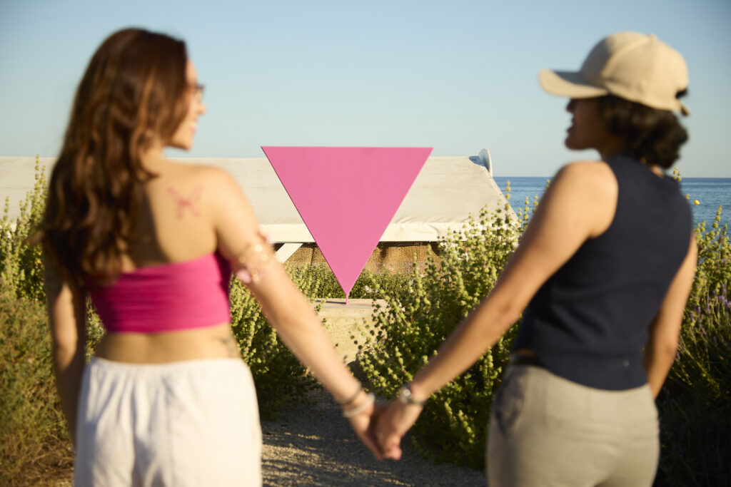 Dues persones agafades de la mà davant d’un triangle rosa, amb el mar de Sitges al fons.
