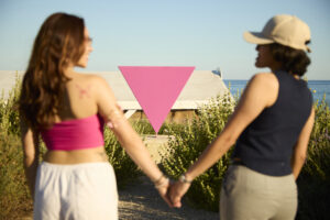 Dues persones agafades de la mà davant d’un triangle rosa, amb el mar de Sitges al fons.