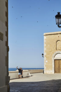 Dues persones fent-se una foto davant del mar, al costat de l’església de Sant Bartomeu i Santa Tecla de Sitges