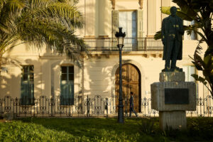 Monument a Santiago Rusiñol davant d’un edifici històric de Sitges, envoltat de vegetació.