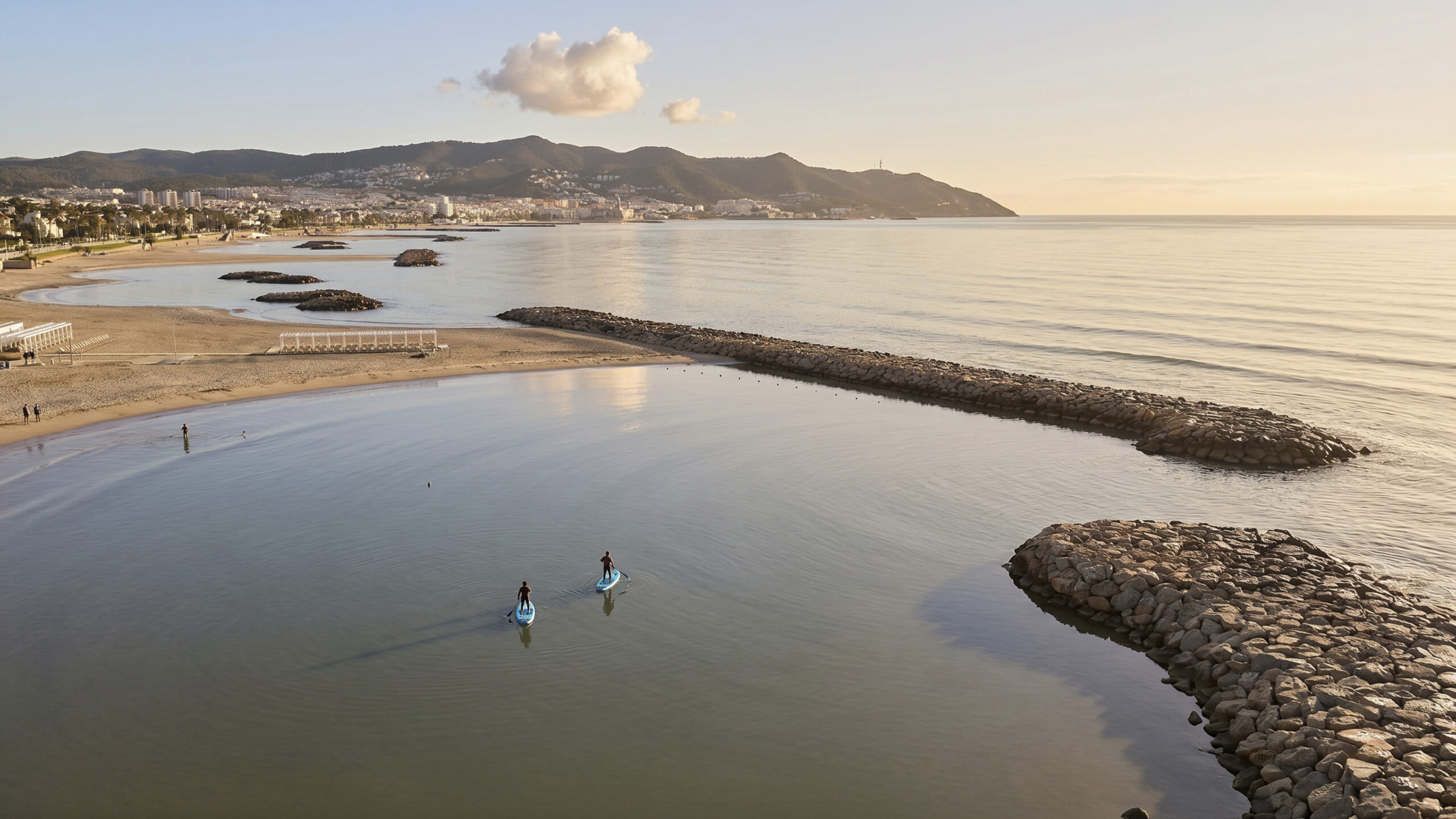 Dues persones practicant pàdel surf davant de les platges de Sitges, amb el massís del Garraf al fons